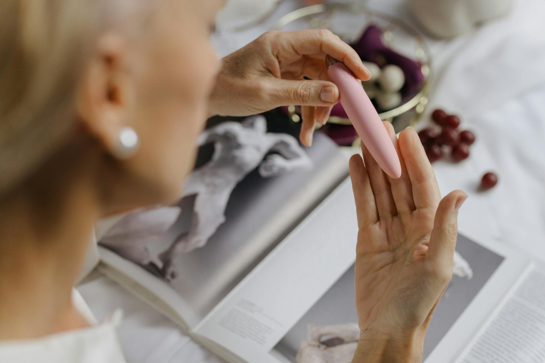 Una mujer sosteniendo un vibrador de color rosa, con libros de arte de fondo, enfatizando temas íntimos y personales.
